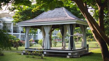 A gazebo with a tiled roof and white walls, surrounded by lush greenery and a well-maintained lawn.