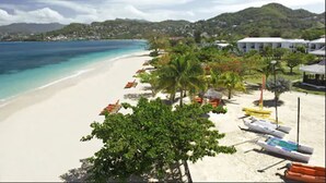 A beach with white sand, clear blue water, and a row of colorful umbrellas. There are palm trees and a building with a white roof in the background. - Coyaba Beach Resort (St. George's)