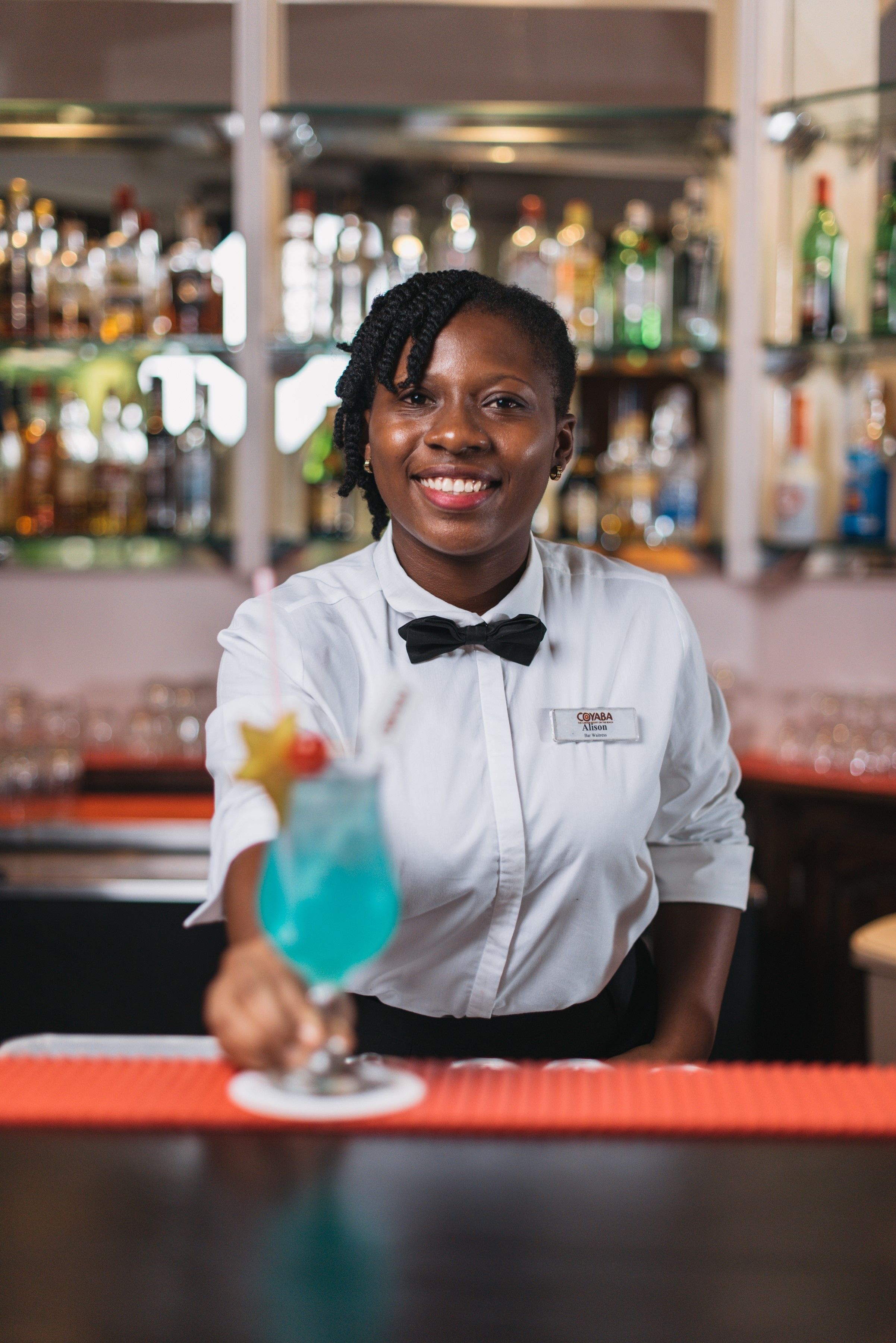 Ein Barkeeper in weißer Hemde und schwarzem Fliege, mit Namensschild, serviert einen blauen Cocktail.