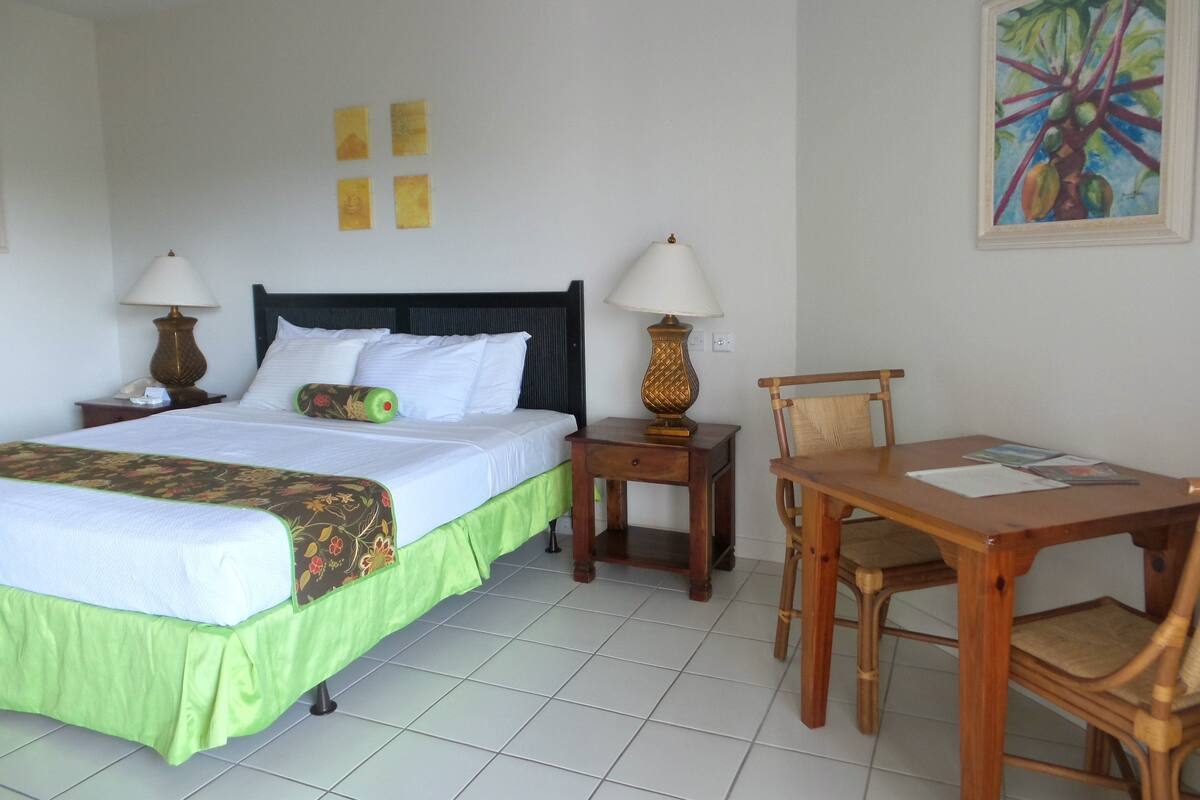 A hotel room with a bed, two bedside tables with lamps, a wooden chair, a small table, and a painting on the wall.