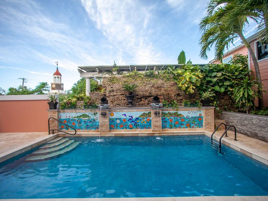 A swimming pool with a tiled mural, a brick wall with plants, and a clock tower in the background.