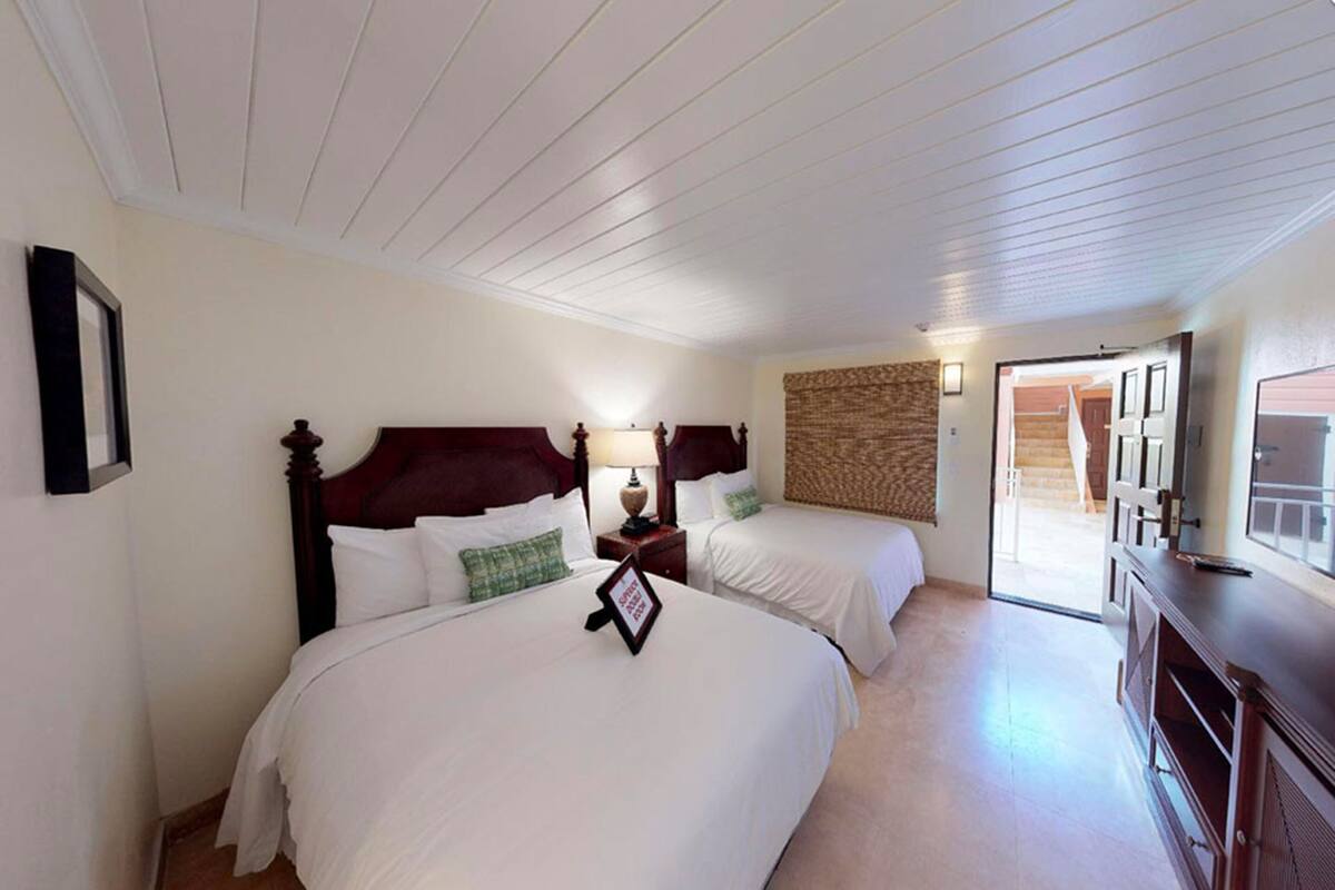 A hotel room with two beds, a wooden headboard, a bedside table with a lamp, a wall-mounted TV, and a door leading to a staircase.