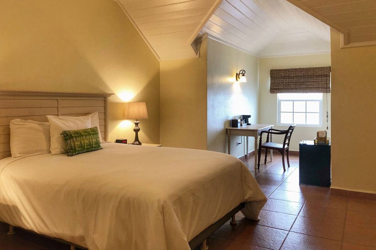 A bedroom with a bed, bedside table, lamp, desk, chair, and a window with blinds.
