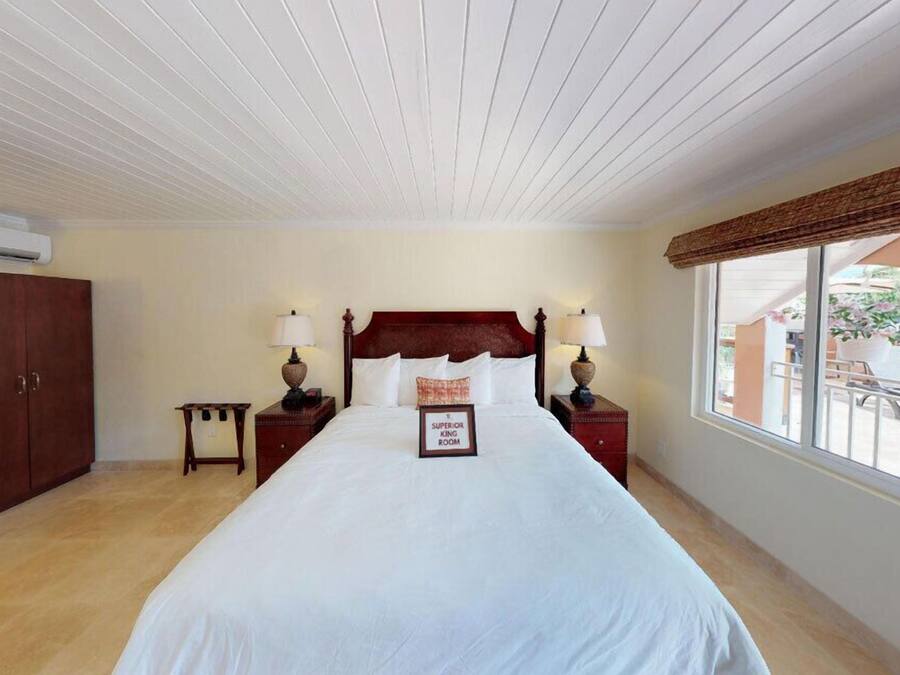 A hotel room with a large bed, wooden headboard, two bedside tables with lamps, a small nightstand, and a window with a view of the outside.