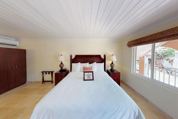 A hotel room with a large bed, wooden headboard, two bedside tables with lamps, a small nightstand, and a window with a view of the outside. - Company House Hotel St. Croix (Christiansted)