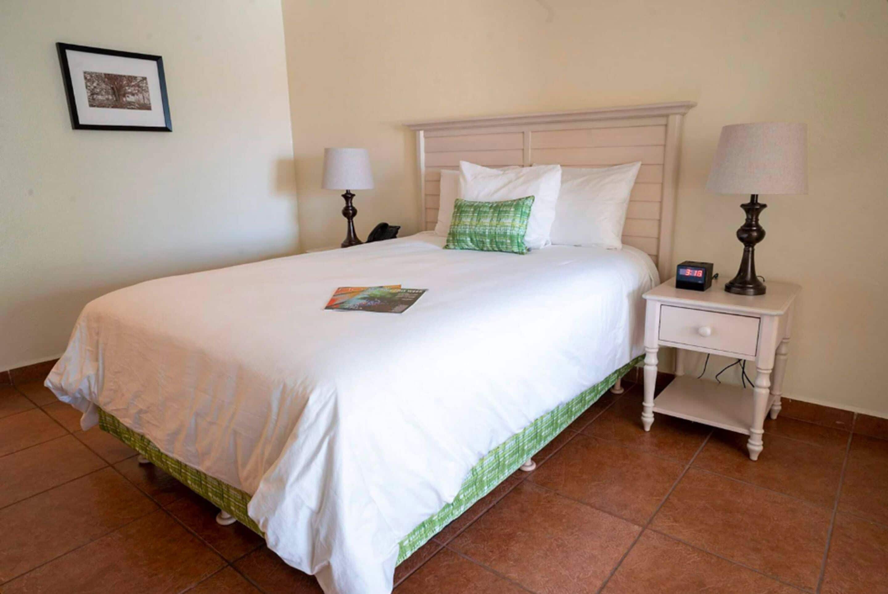 Una cama bien hecha con ropa de cama blanca y un cojín verde decorativo, una mesita de noche con lámpara y un cuadro en la pared.