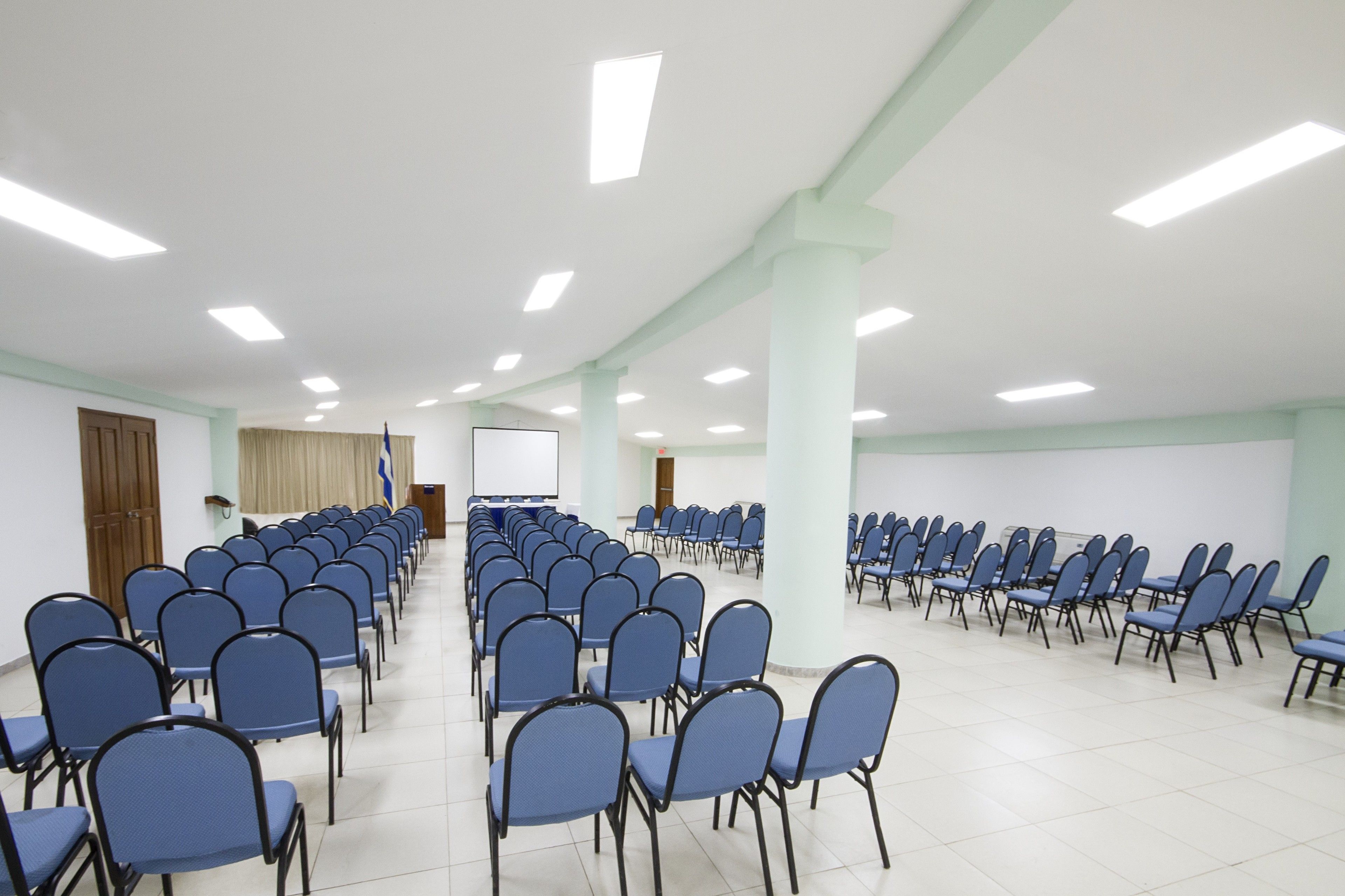 Uma sala de conferências com fileiras de cadeiras azuis, um quadro branco e uma tela de projetor.