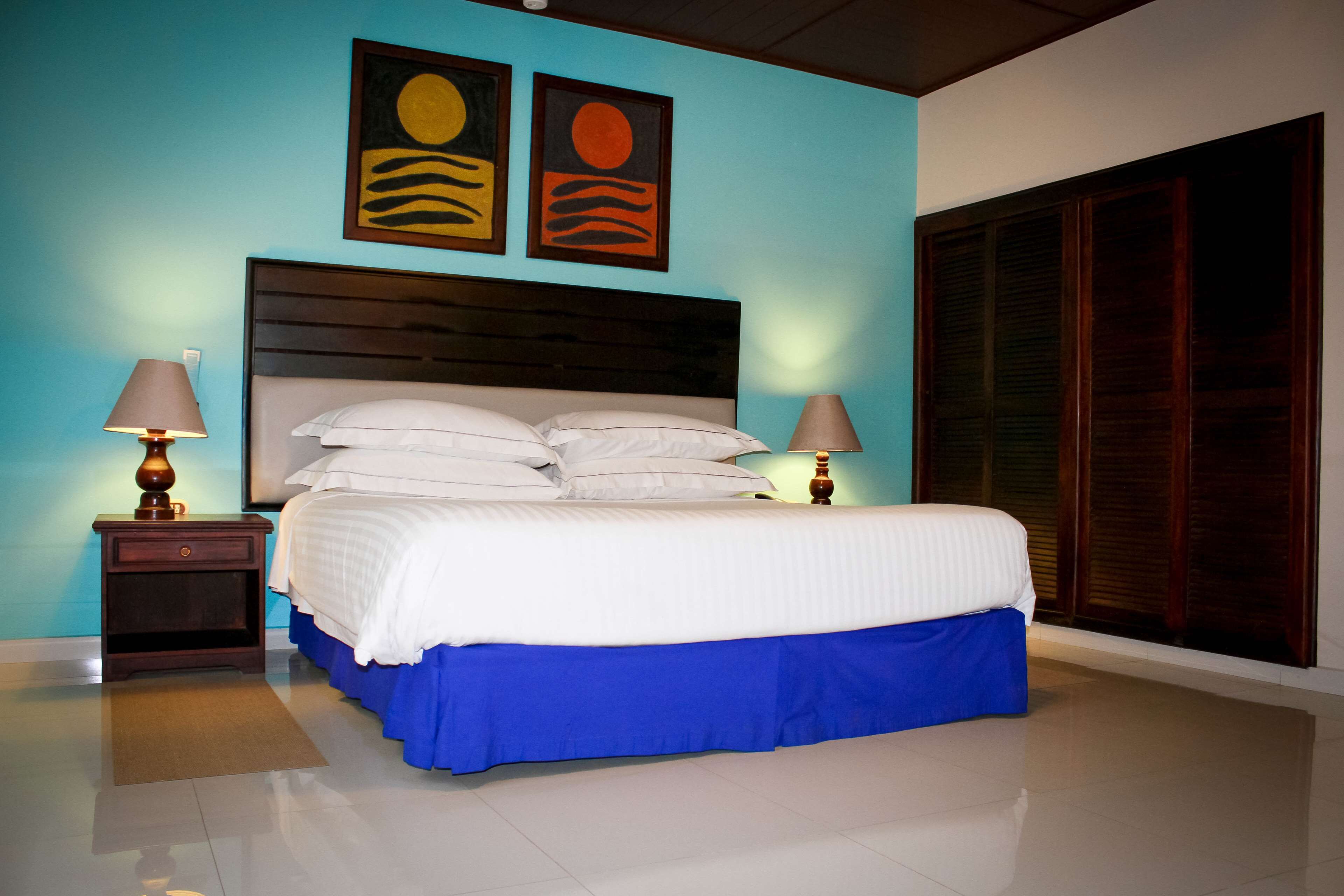 Une chambre à coucher comprenant un lit, deux tables de chevet, un sommier de lit bleu avec une jupe, deux lampes fixées au mur et deux tableaux au-dessus du lit.