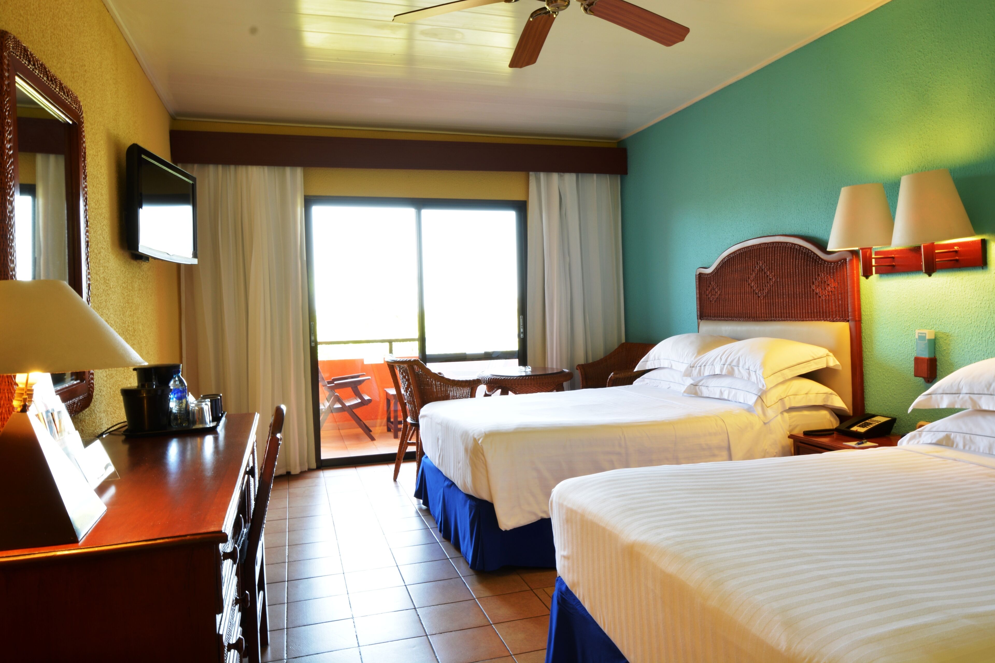 A hotel room with two beds, a desk, a chair, a television, and a ceiling fan.