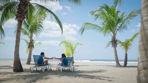 Dos personas sentadas en sillas de playa, mirándose, con palmeras y el océano al fondo.