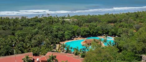 A resort with a large pool, surrounded by dense tropical vegetation and a view of the ocean.
