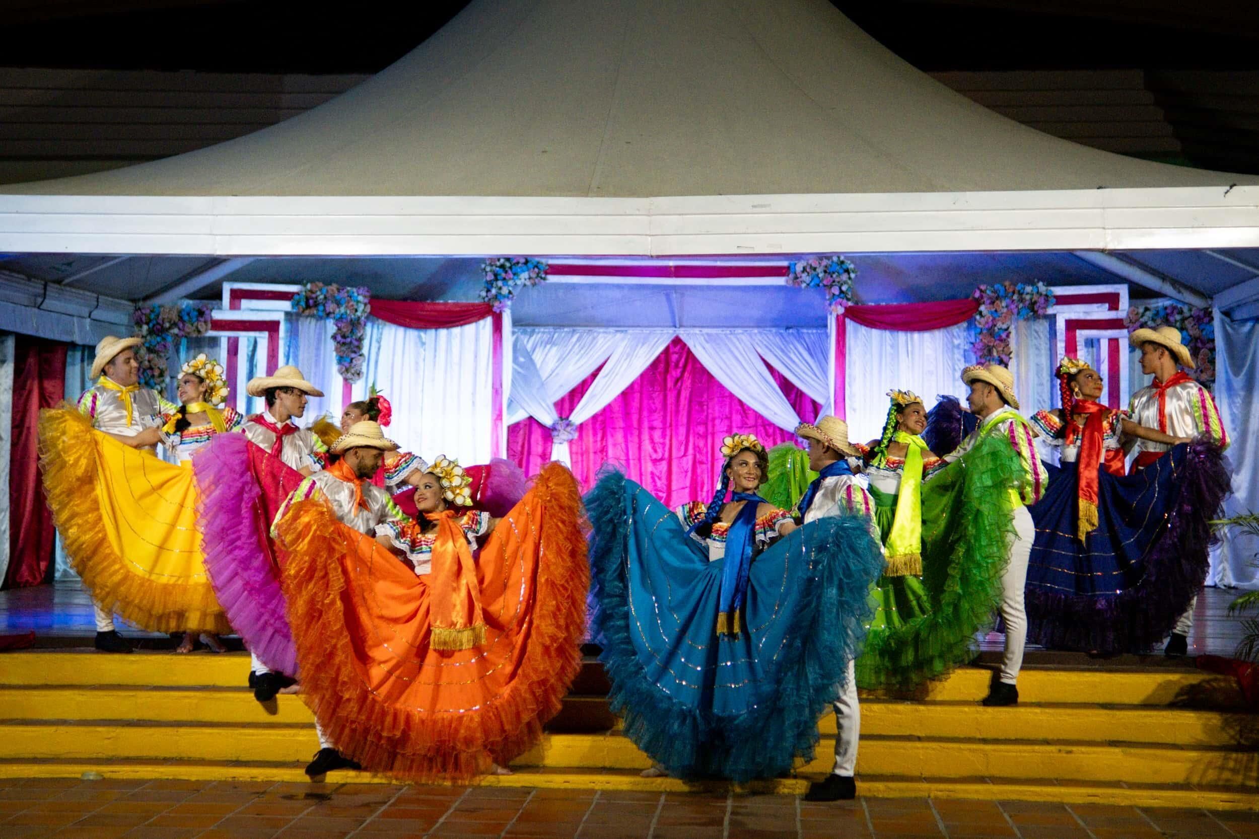 A group of people in colorful traditional costumes performing on a stage with a white tent and pink curtains.