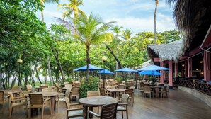 An outdoor seating area with round tables and chairs, surrounded by palm trees and a thatched-roof structure. - Barceló Montelimar - All Inclusive (San Rafael del Sur)