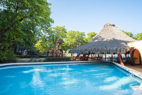 Una piscina con una estructura de techo de paja, rodeada de follaje y un tobogán.