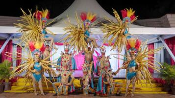 A group of performers in elaborate costumes and feathered headdresses on a stage with a white tent backdrop.