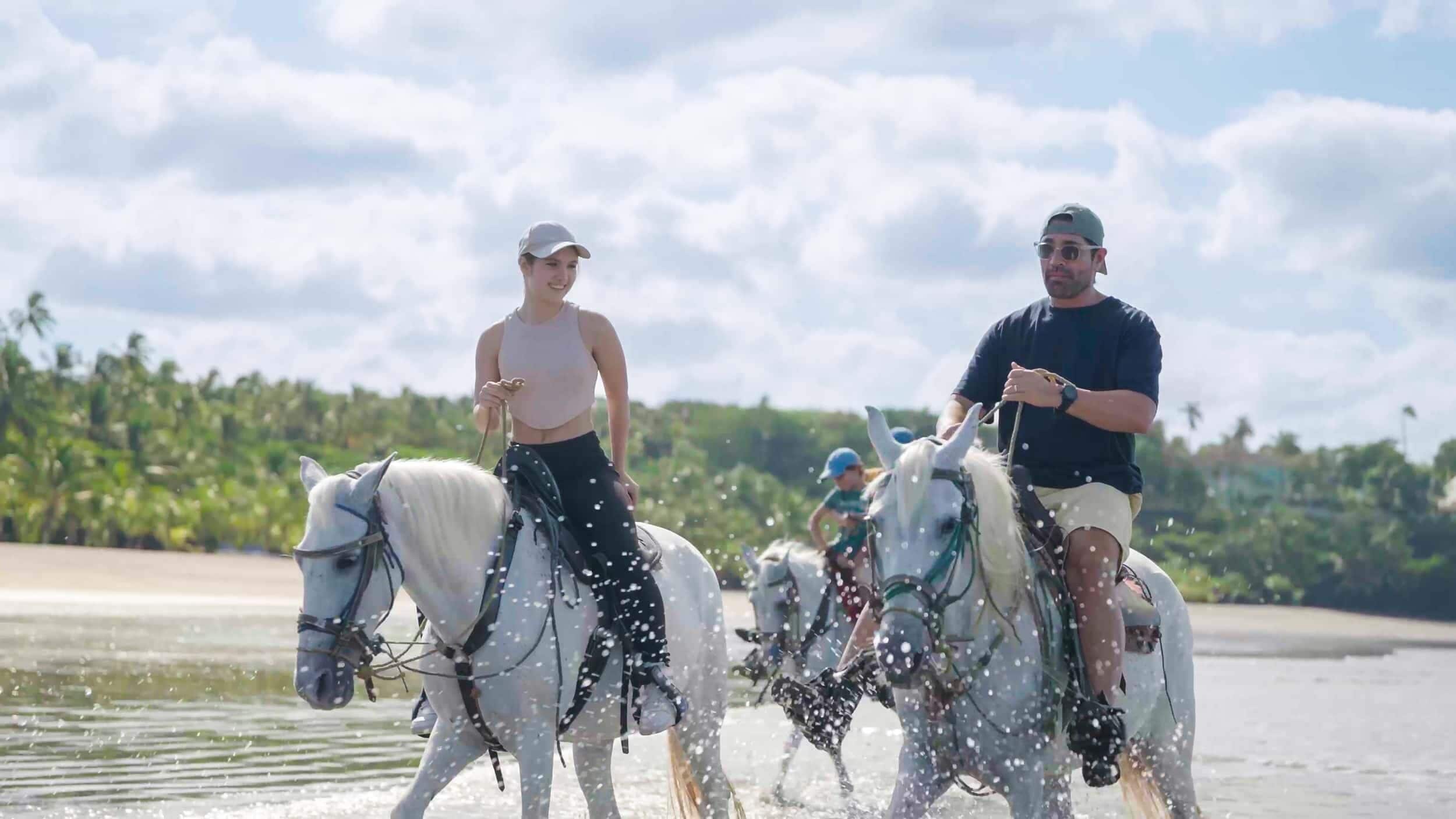 Two people riding horses along a water body with a tropical background.