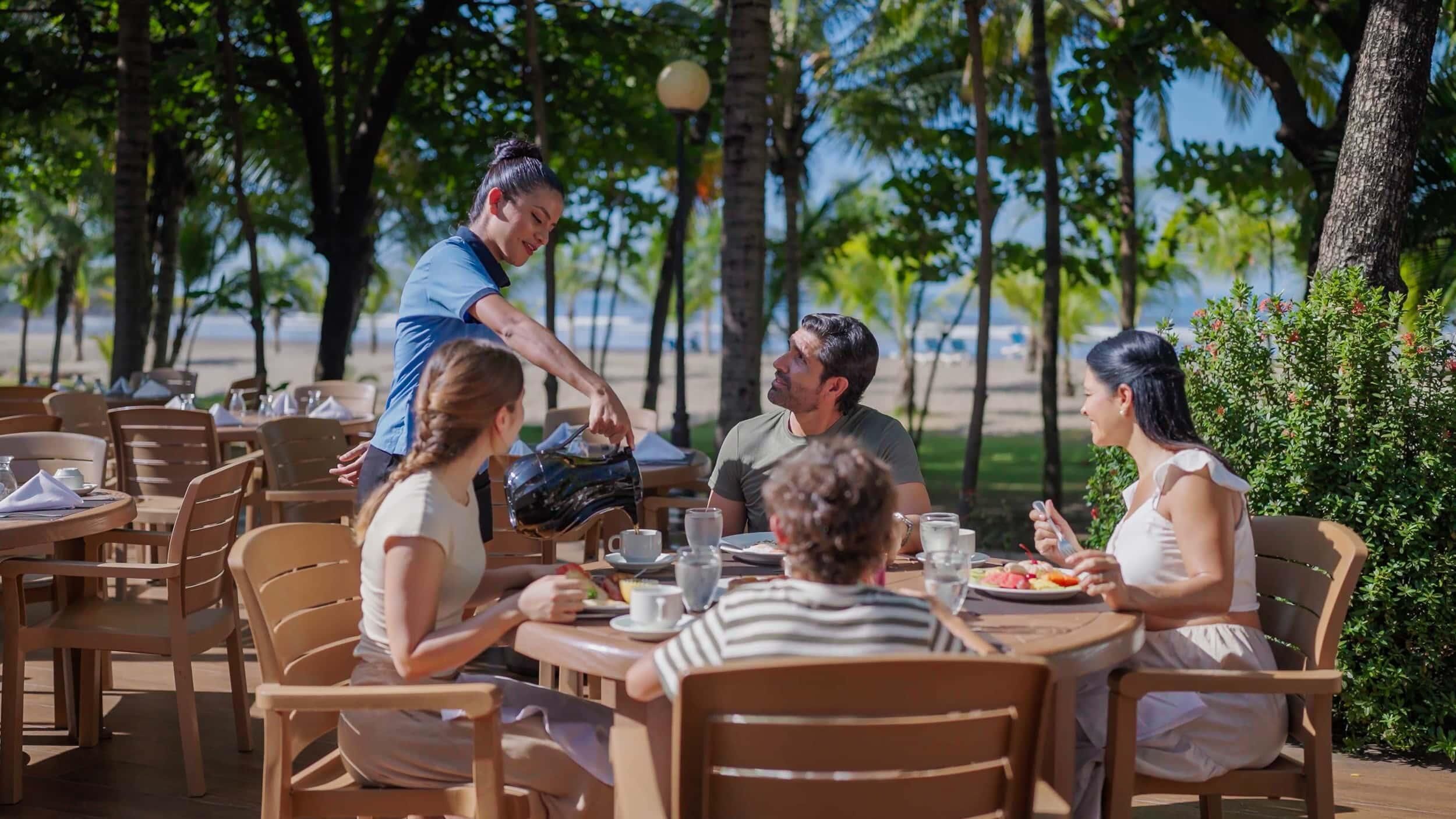 An outdoor dining area with wooden tables and chairs, surrounded by palm trees and a view of the beach.