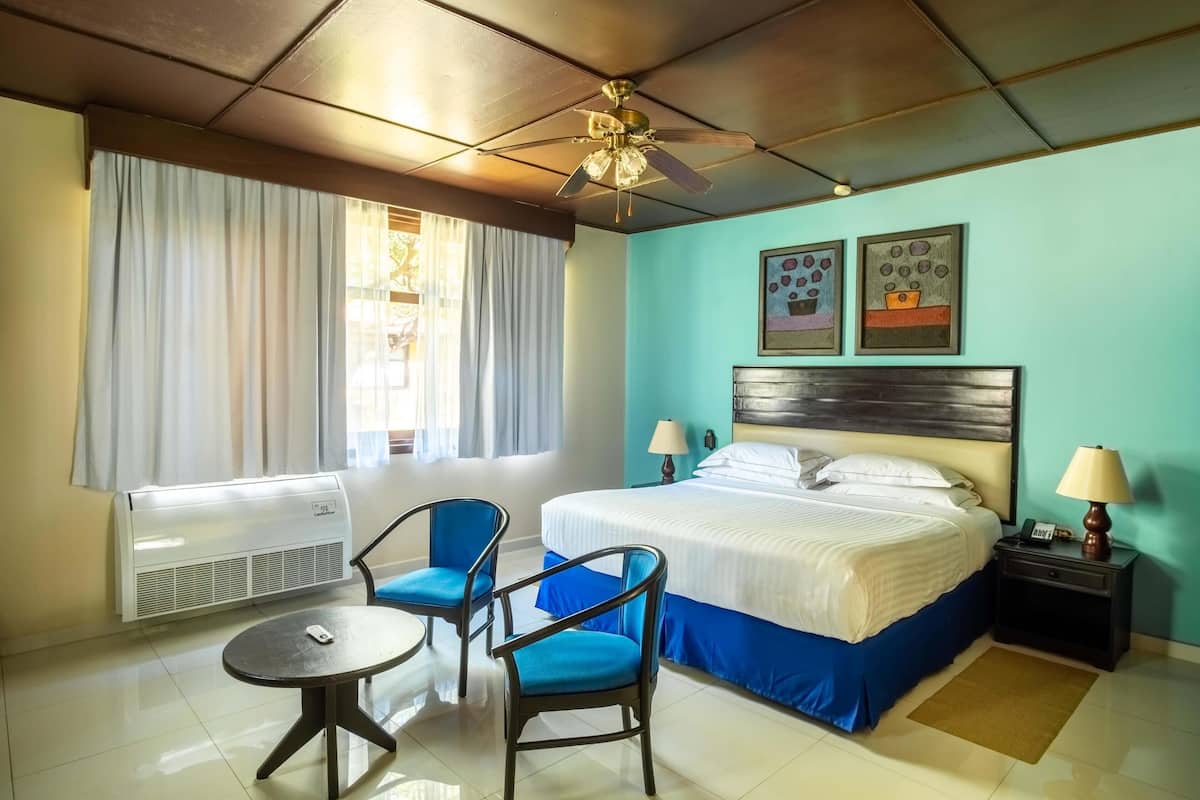 A hotel room with a bed, two chairs, a table, a ceiling fan, a window with curtains, and a wall-mounted air conditioner.