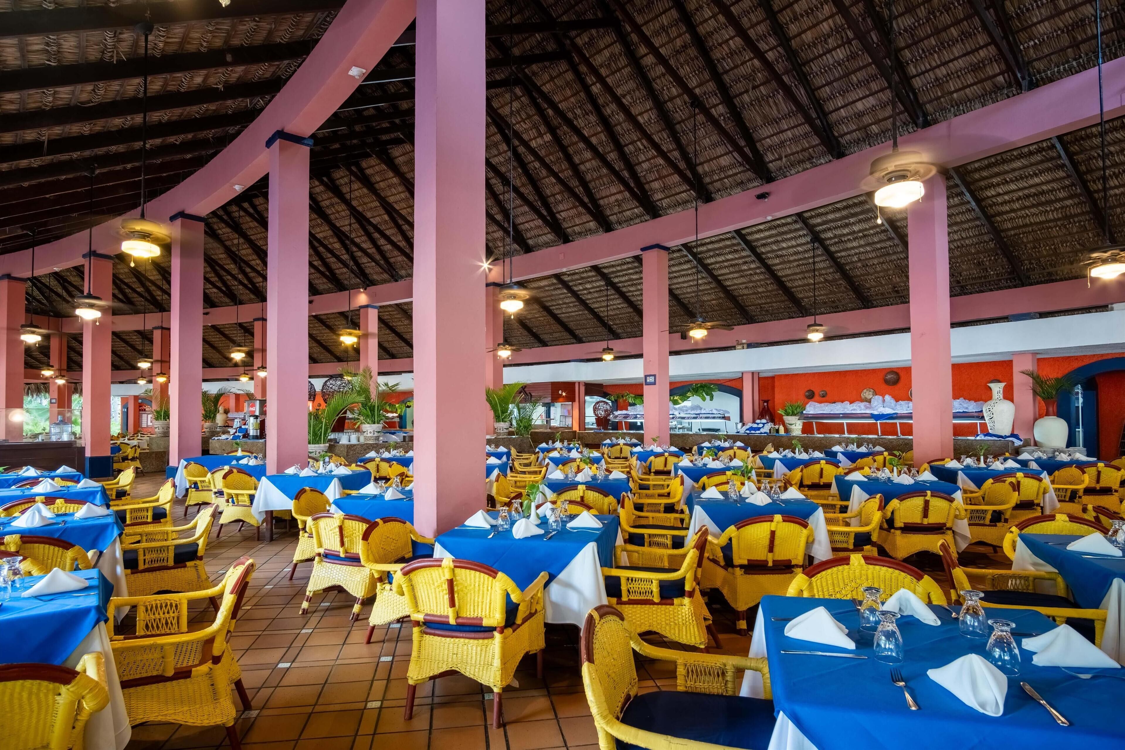 A large indoor dining area with tables set for multiple guests, featuring yellow wicker chairs and blue tablecloths.
