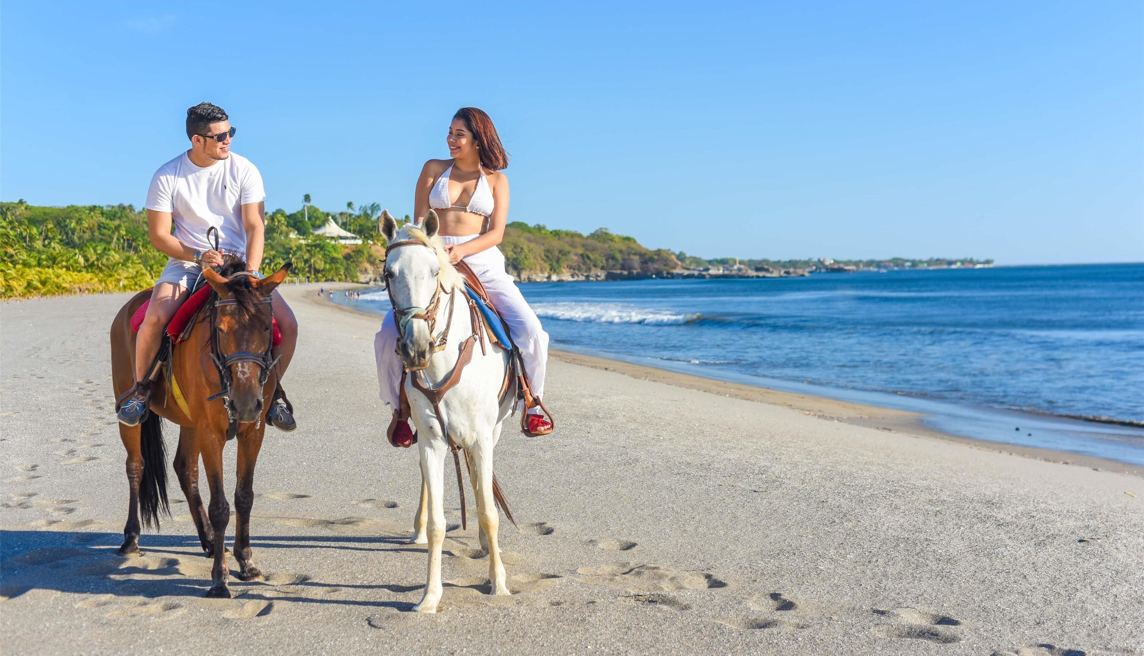 Deux personnes à cheval sur une plage, avec une vue dégagée sur l’océan et une côte lointaine.