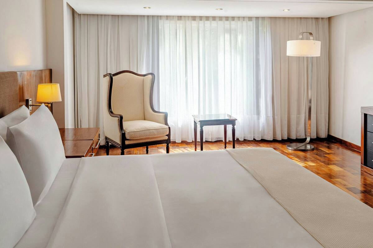 Habitación de hotel con una cama grande, una silla, una mesita, una lámpara y vistas a través de cortinas translúcidas.