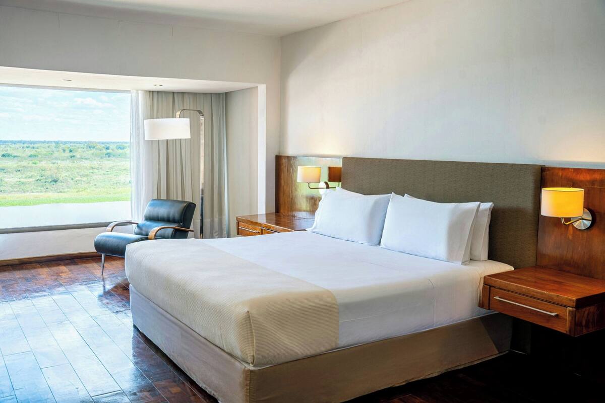 Habitación de hotel con una cama grande, una silla, una mesita de noche y vistas al exterior.