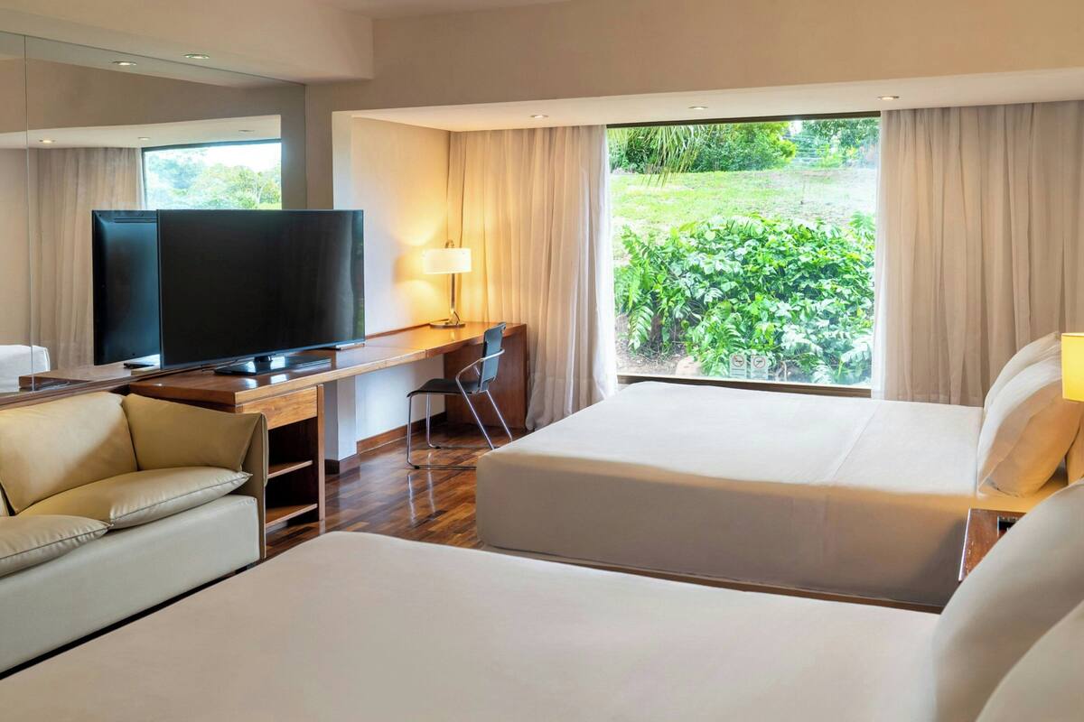 Habitación de hotel con cama, escritorio con televisor, sofá y vista a zonas verdes.