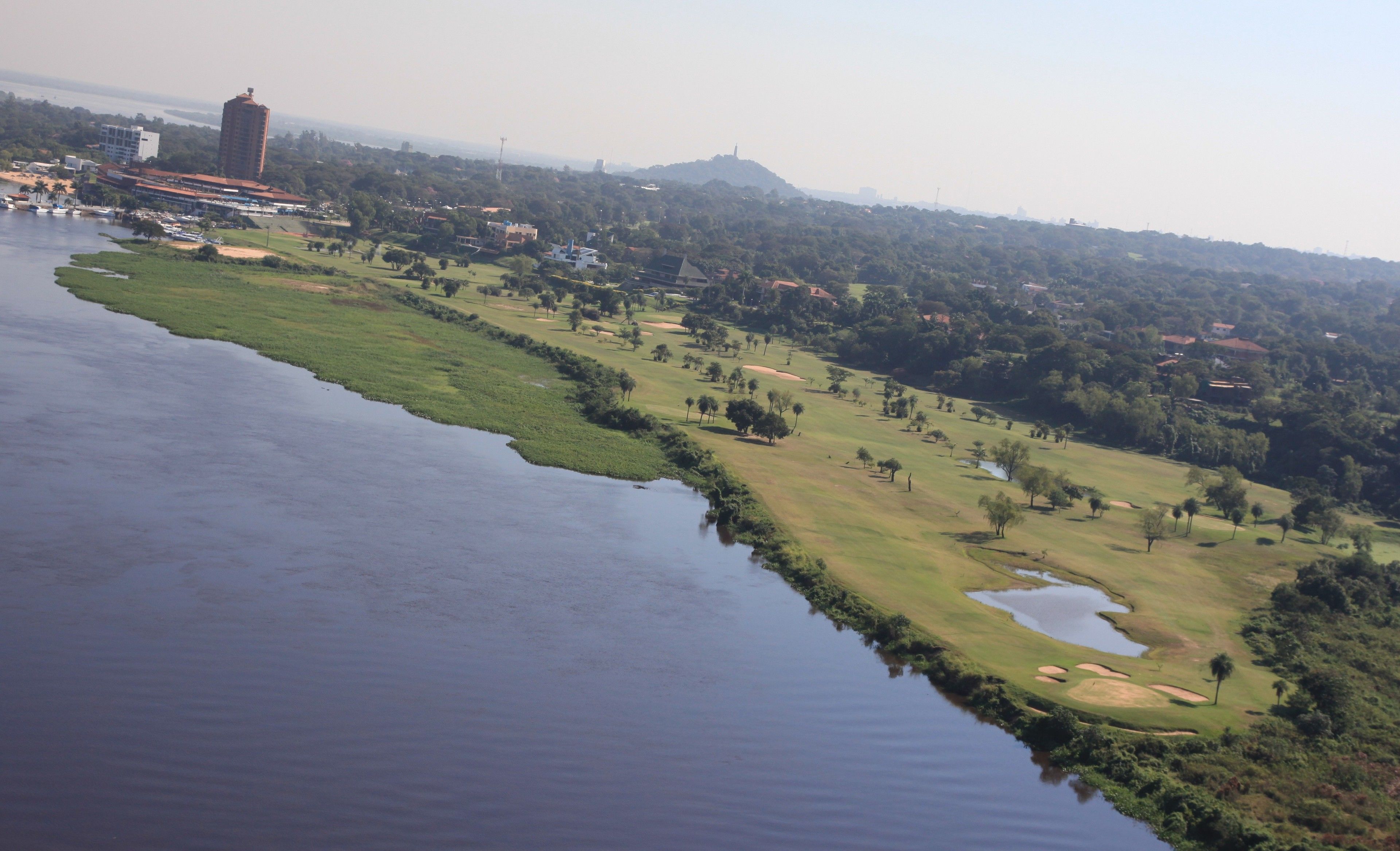 Vista aérea de un campo de golf con una masa de agua a un lado, una colina con un monumento a lo lejos y edificios al fondo.
