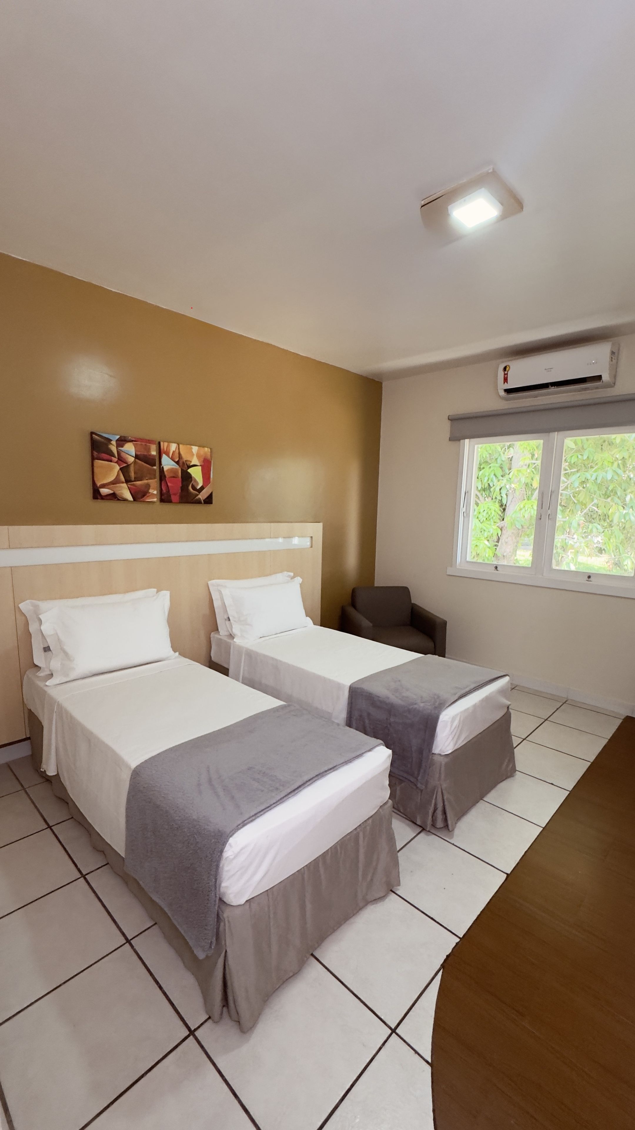 A hotel room with two beds, a window with a view of greenery, and an air conditioning unit.