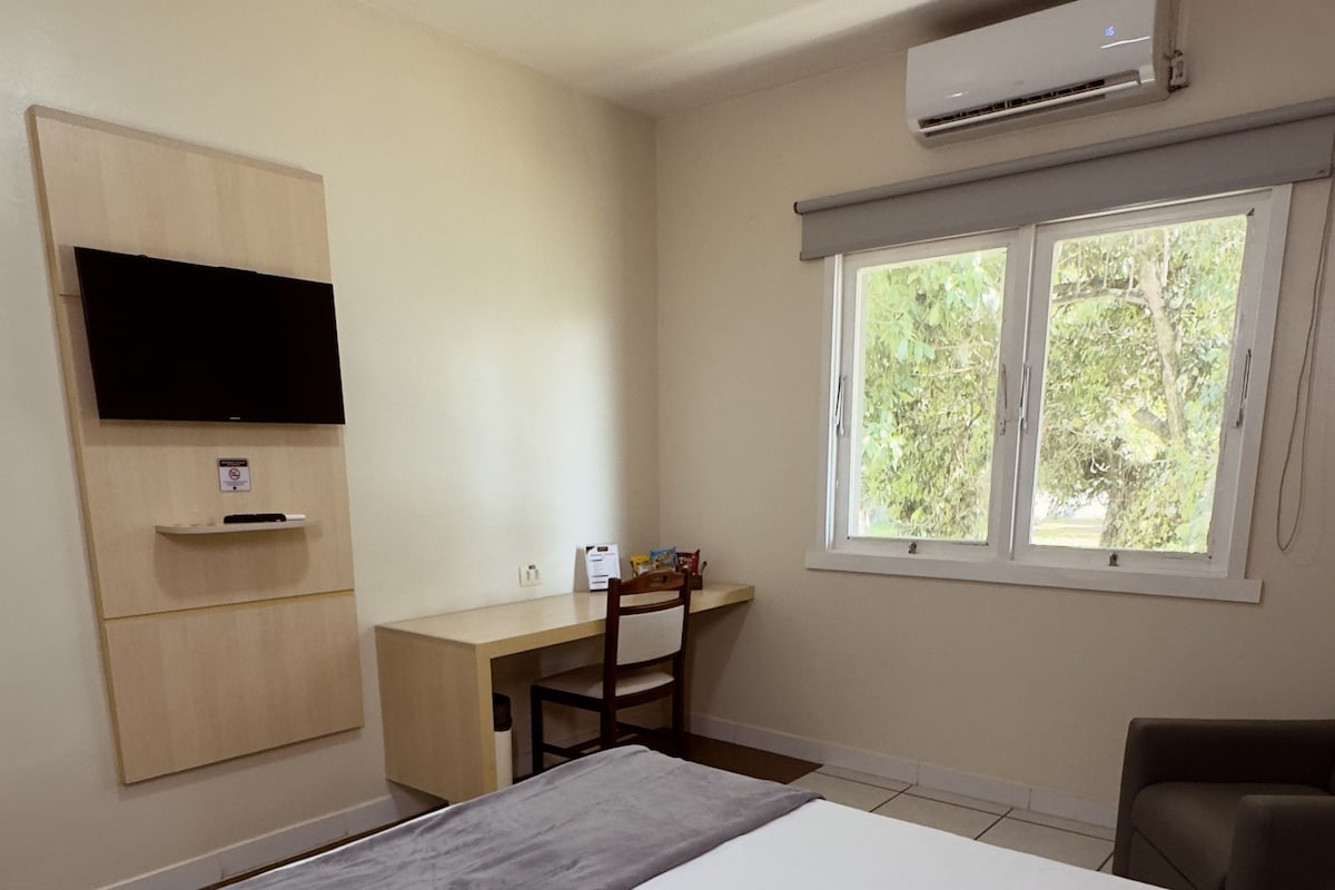 Un dormitorio con cama, escritorio, silla, televisión y ventana con vista a follaje.