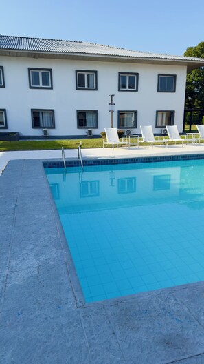 Una piscina con superficie de agua azul cristalina, rodeada por una solana de piedra. Hay sillas reclinables blancas y un edificio con múltiples ventanas al fondo.