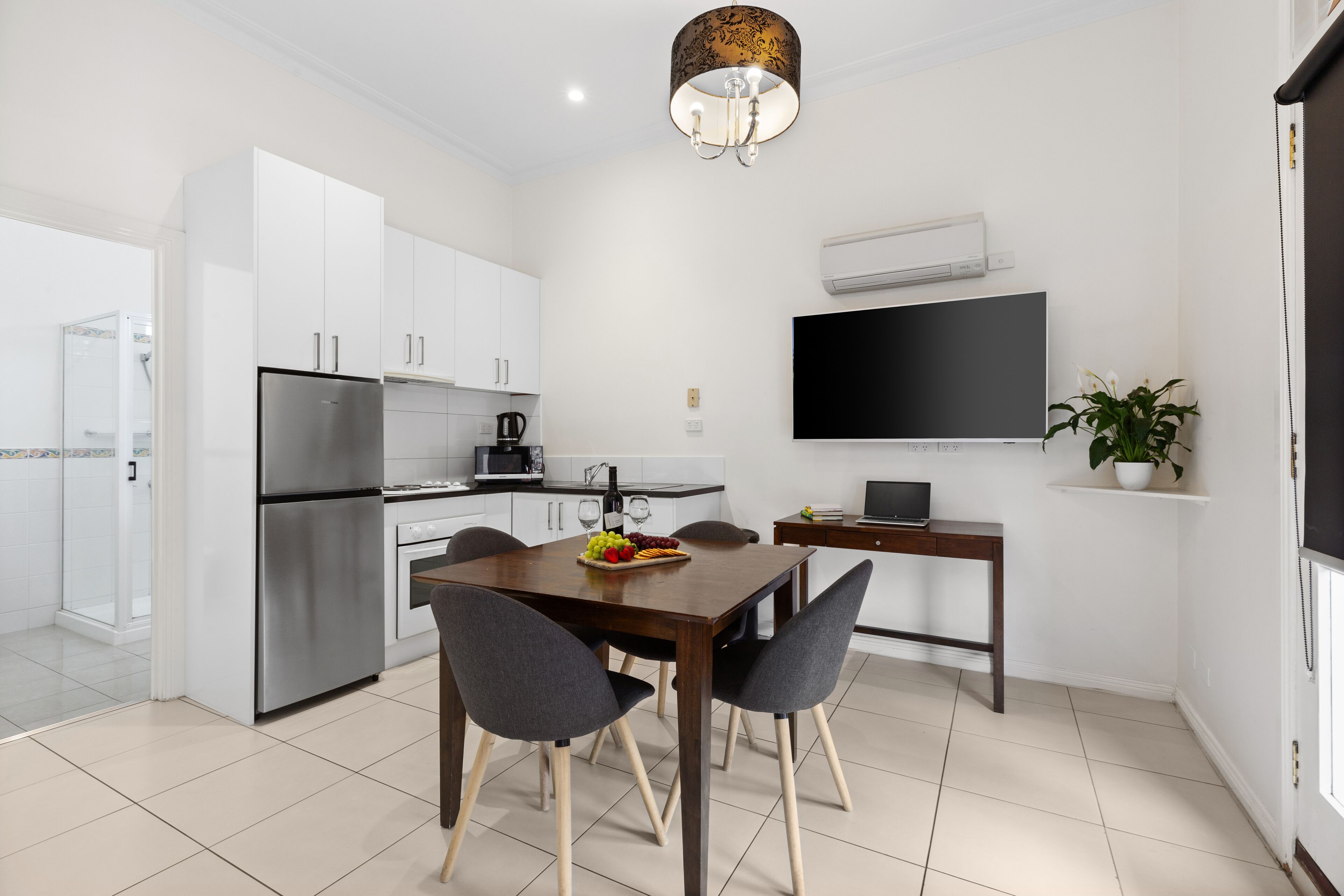 A modern kitchen with white cabinets, a stainless steel refrigerator, a dining table with four chairs, and a flat-screen TV mounted on the wall.