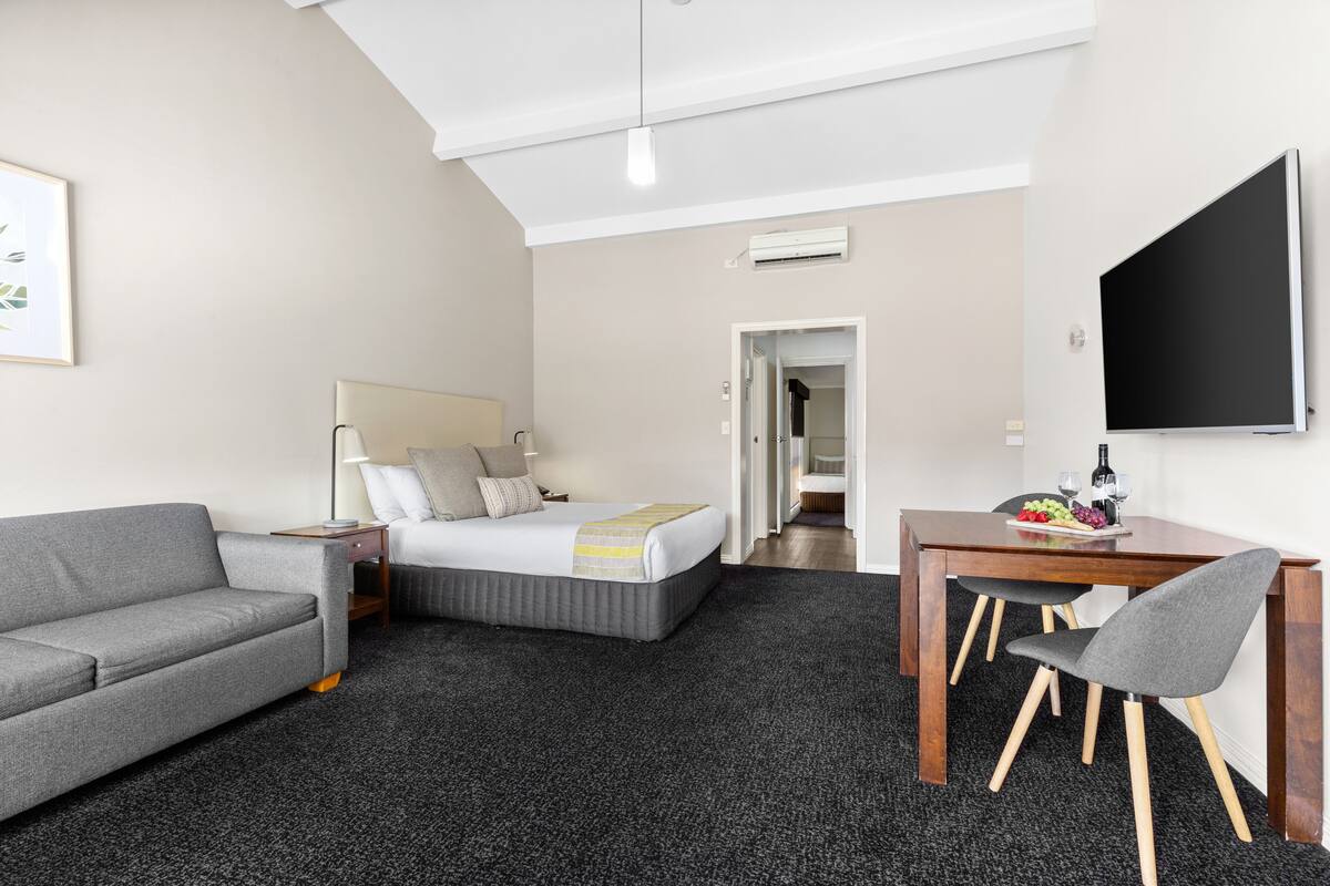 Quarto de hotel com cama, sofá cinzento, mesa de jantar de madeira com duas cadeiras e televisão de tela plana.