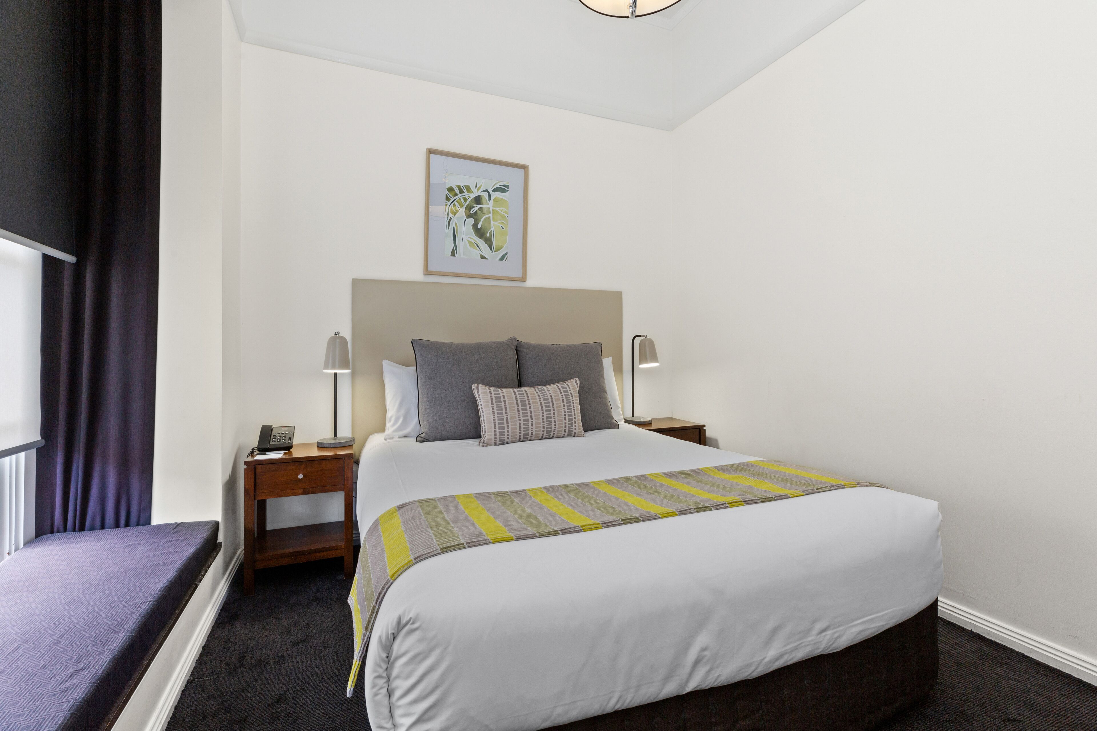Ein Hotelzimmer mit einem großen Bett, zwei Nachttischen mit Lampen, einem kleinen Nachttisch mit Telefon, einem Fenster mit Vorhängen und einem gerahmten Bild an der Wand.
