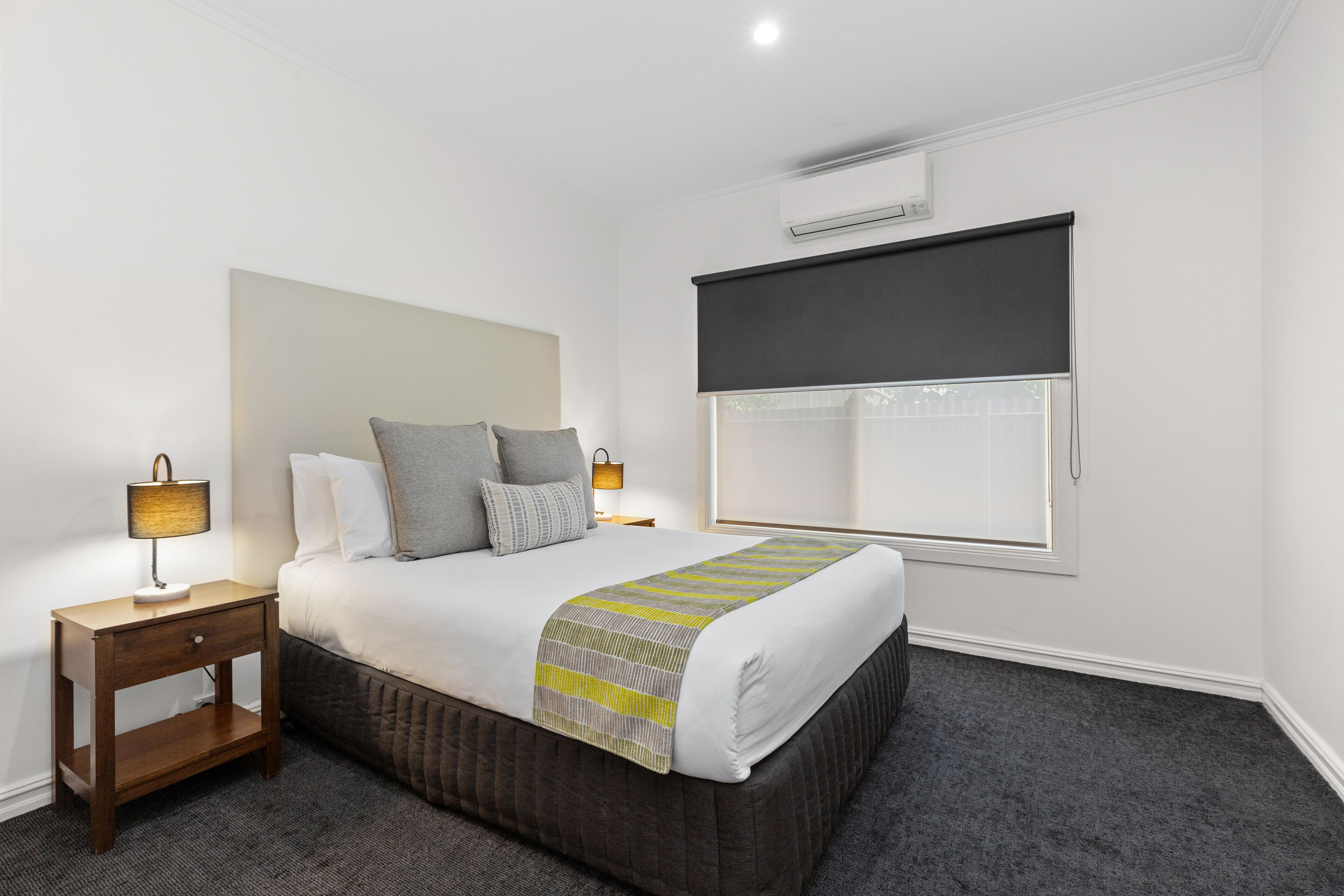 Une chambre à coucher bien rangée, avec un lit, des lampes de chevet, une fenêtre avec un store et un appareil de climatisation fixé au mur.