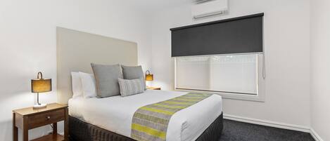 A neatly arranged bedroom with a bed, bedside lamps, a window with a blind, and an air conditioning unit on the wall.