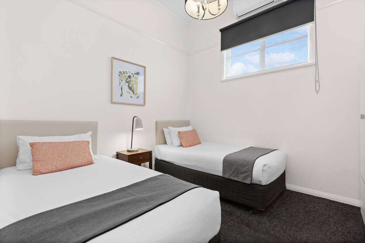 Quarto de hotel com duas camas, uma mesa de cabeceira, uma luminária, um quadro pendurado na parede e uma janela com vista para o céu.