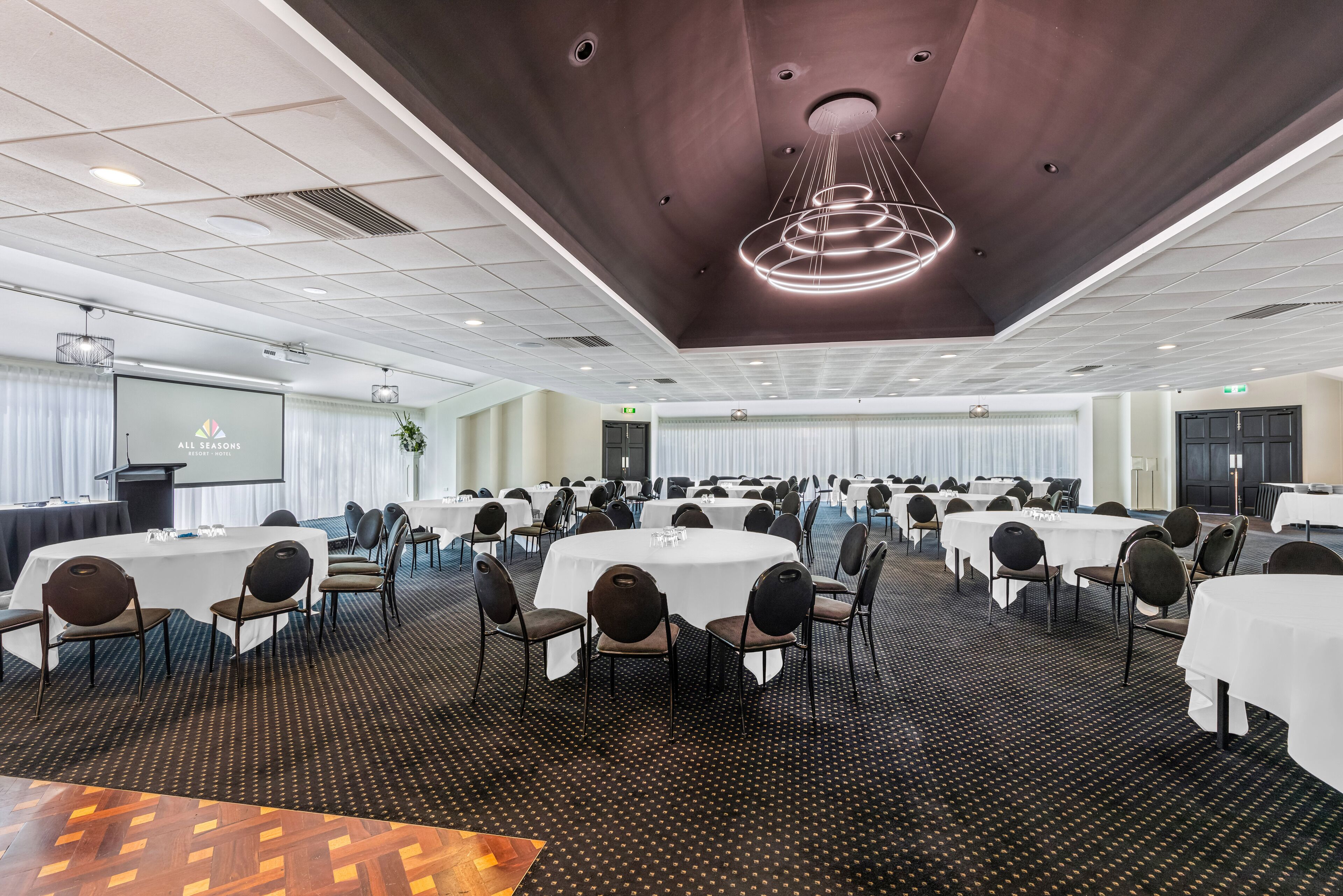 Un salón de conferencias con varias mesas redondas y sillas, una pantalla grande y un diseño de techo moderno.