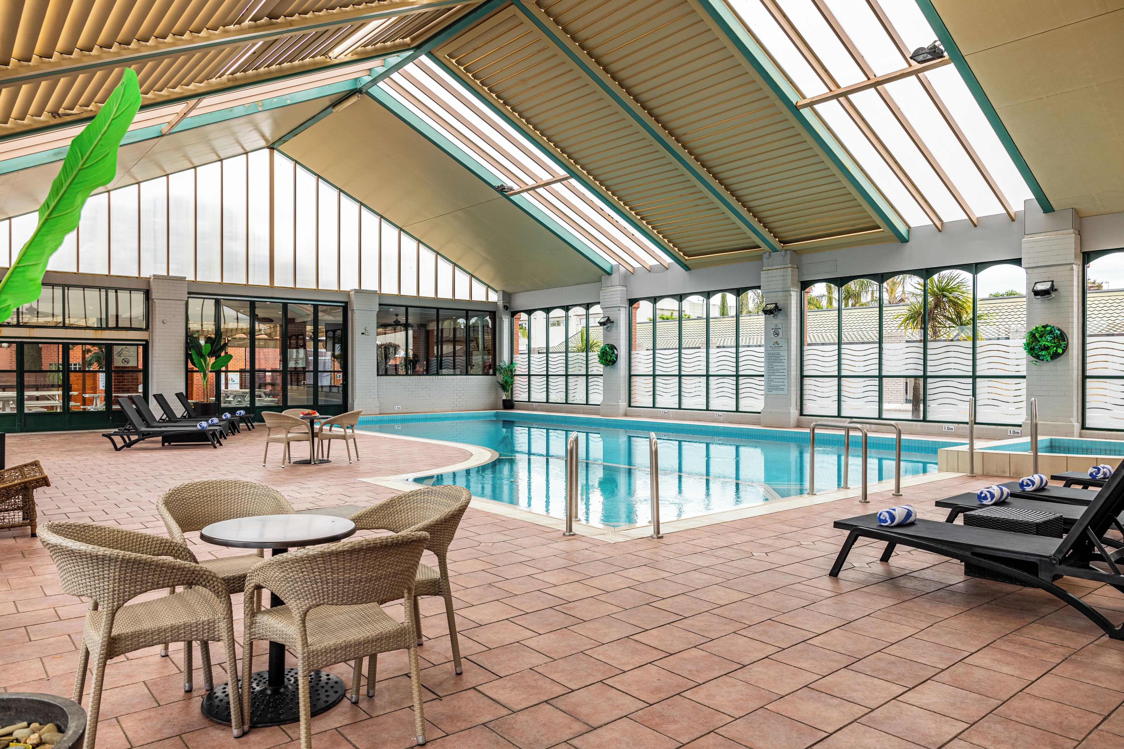 An indoor swimming pool with lounge chairs and tables, surrounded by large windows and a high ceiling.