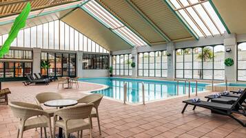 An indoor swimming pool with lounge chairs and tables, surrounded by large windows and a high ceiling.