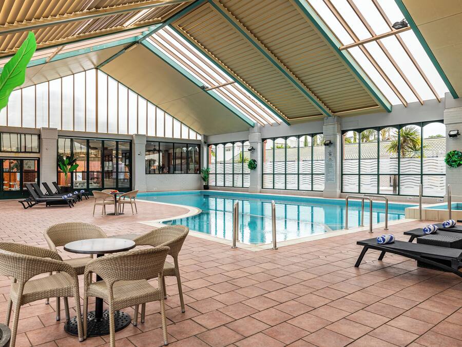 Ein Hallenbad mit Liegestühlen und Tischen, umgeben von großen Fenstern und einer hohen Decke.
