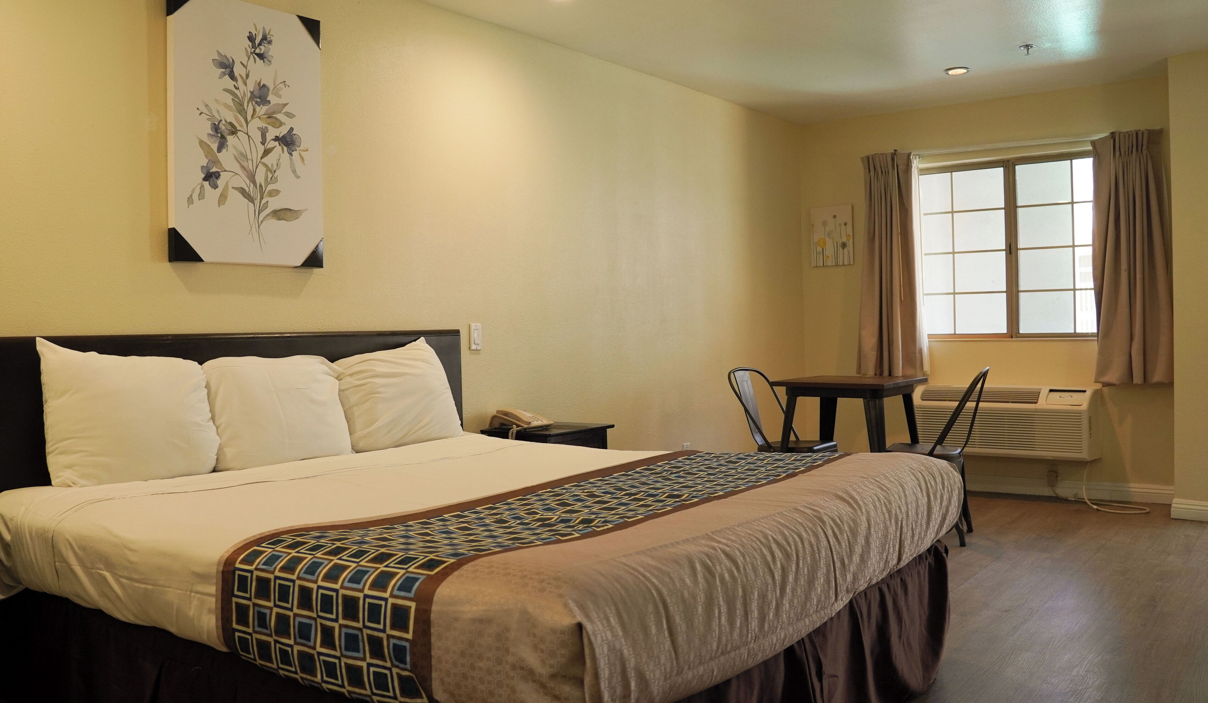 A hotel room with a bed, a desk, a chair, a window with curtains, and an air conditioning unit.