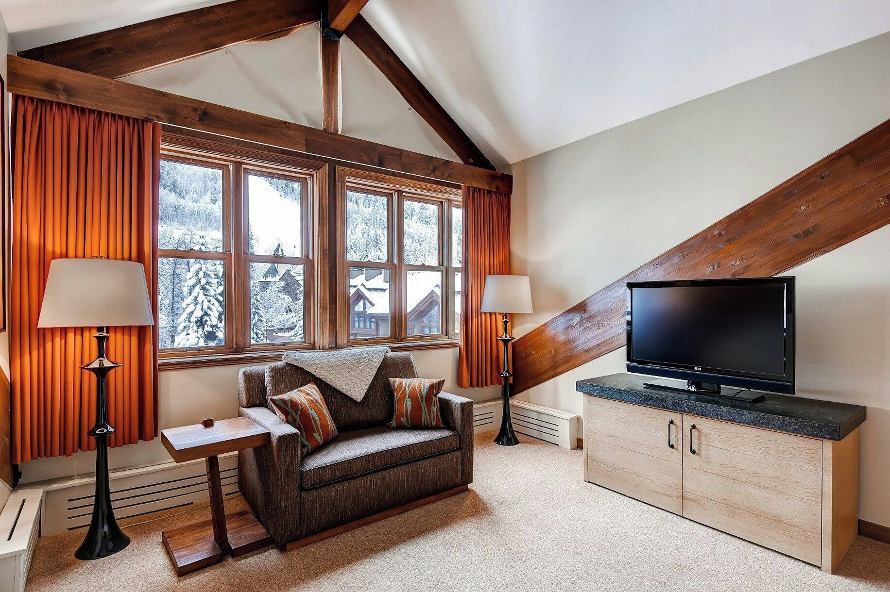A cozy room with a wooden ceiling, a sofa with cushions, a television on a stand, and a window with curtains.