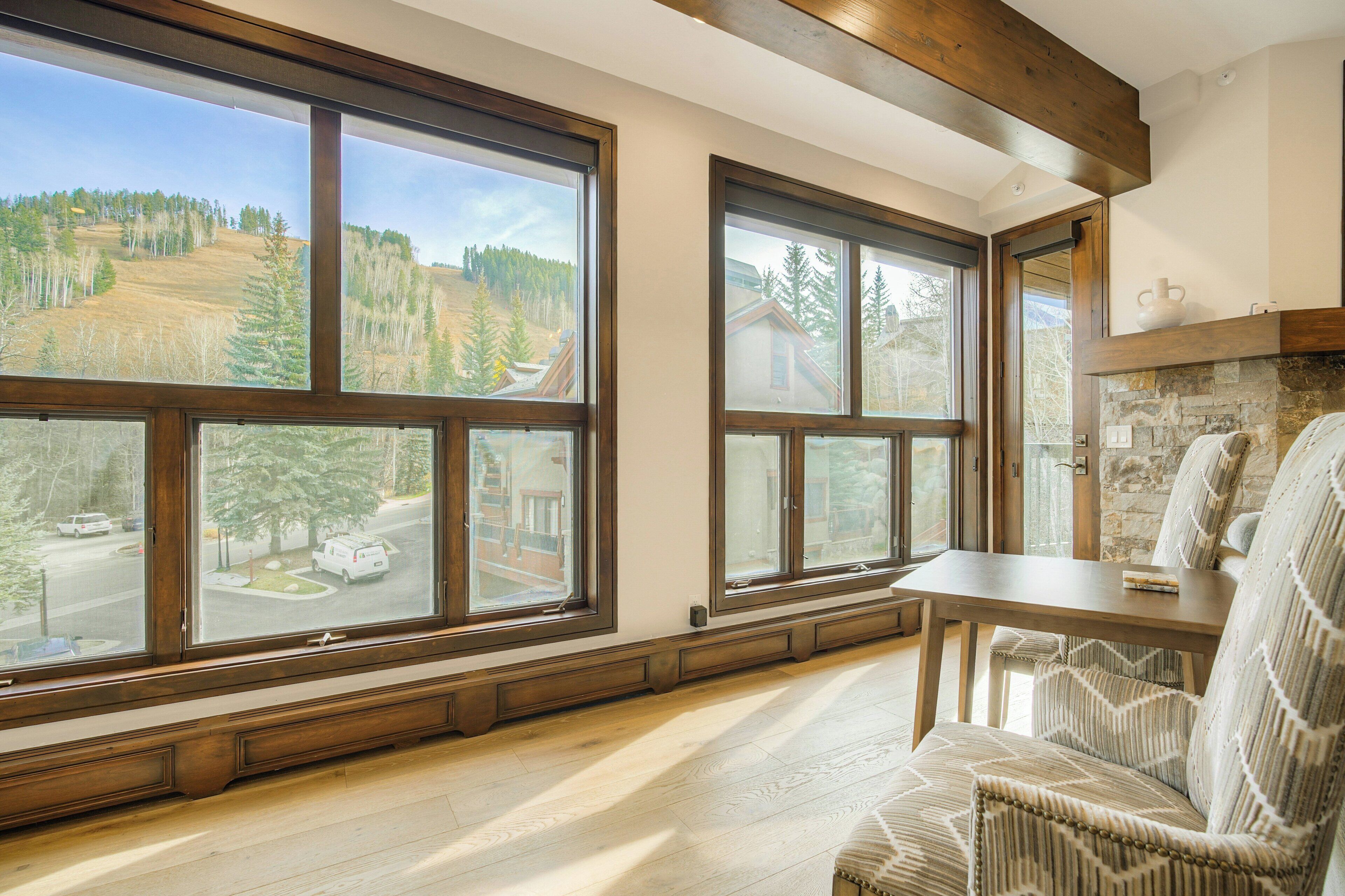 A room with large windows offering a view of a forested hillside. There is a wooden table and a cushioned chair.