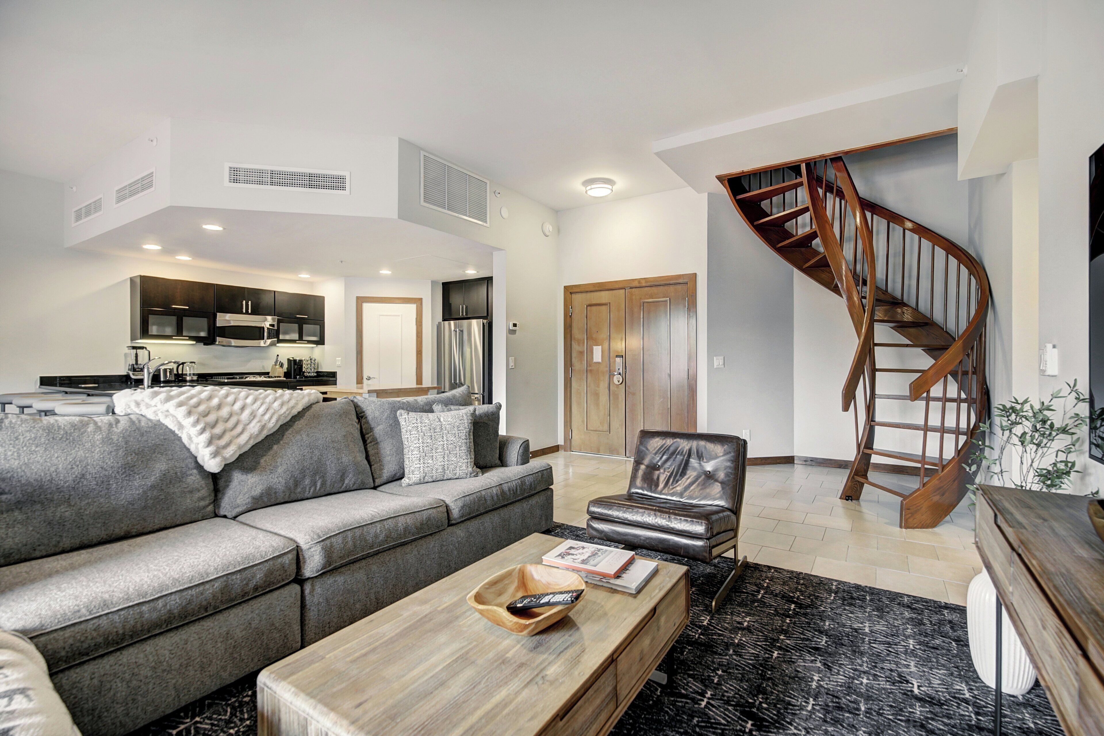 Un salon moderne avec un canapé gris, un fauteuil en cuir, une table basse en bois, un tapis noir et blanc et un escalier en bois.