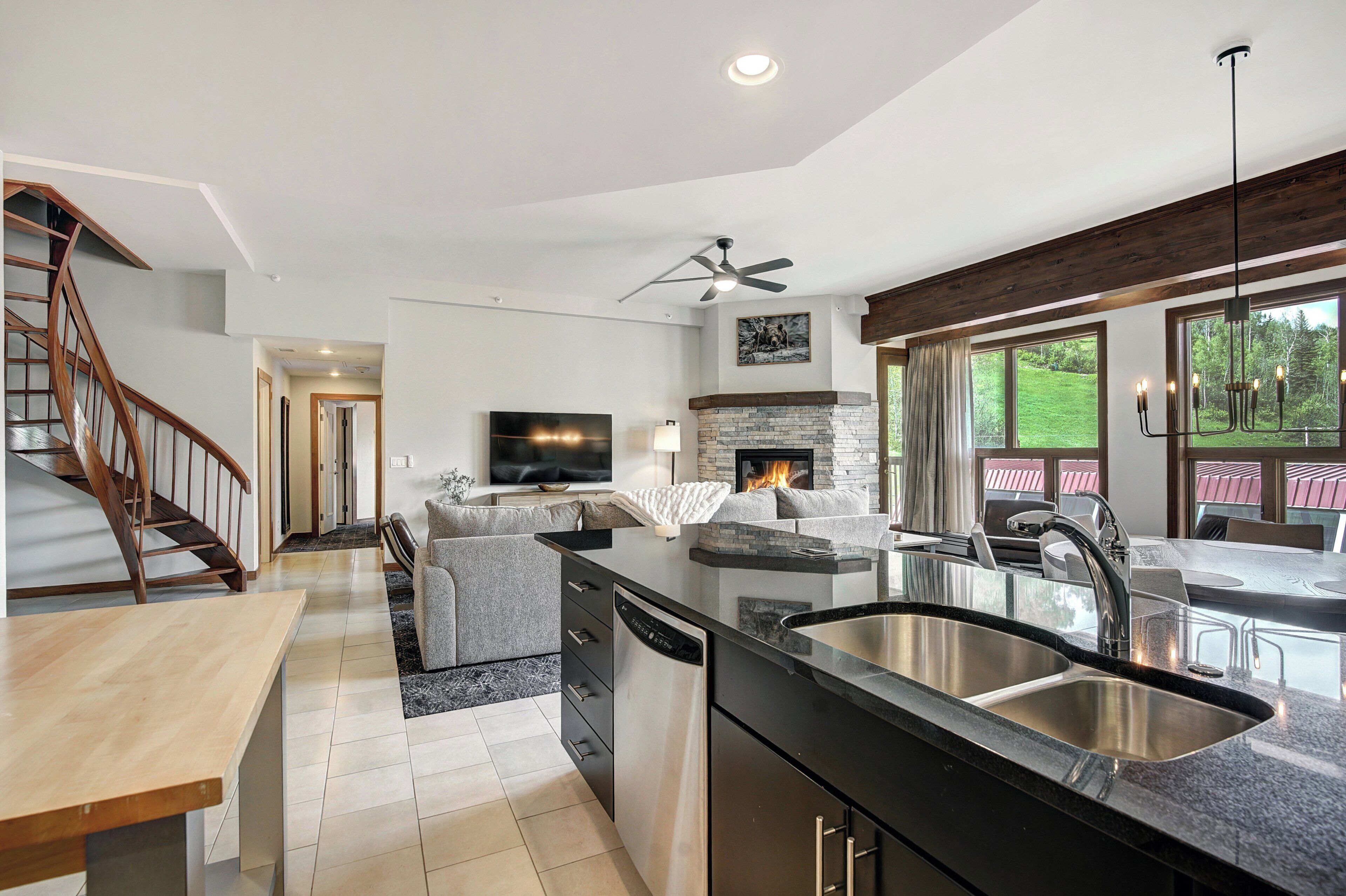 A modern kitchen with a central island, a dining area with a wooden table, a built-in fireplace, and a large window overlooking a green landscape.