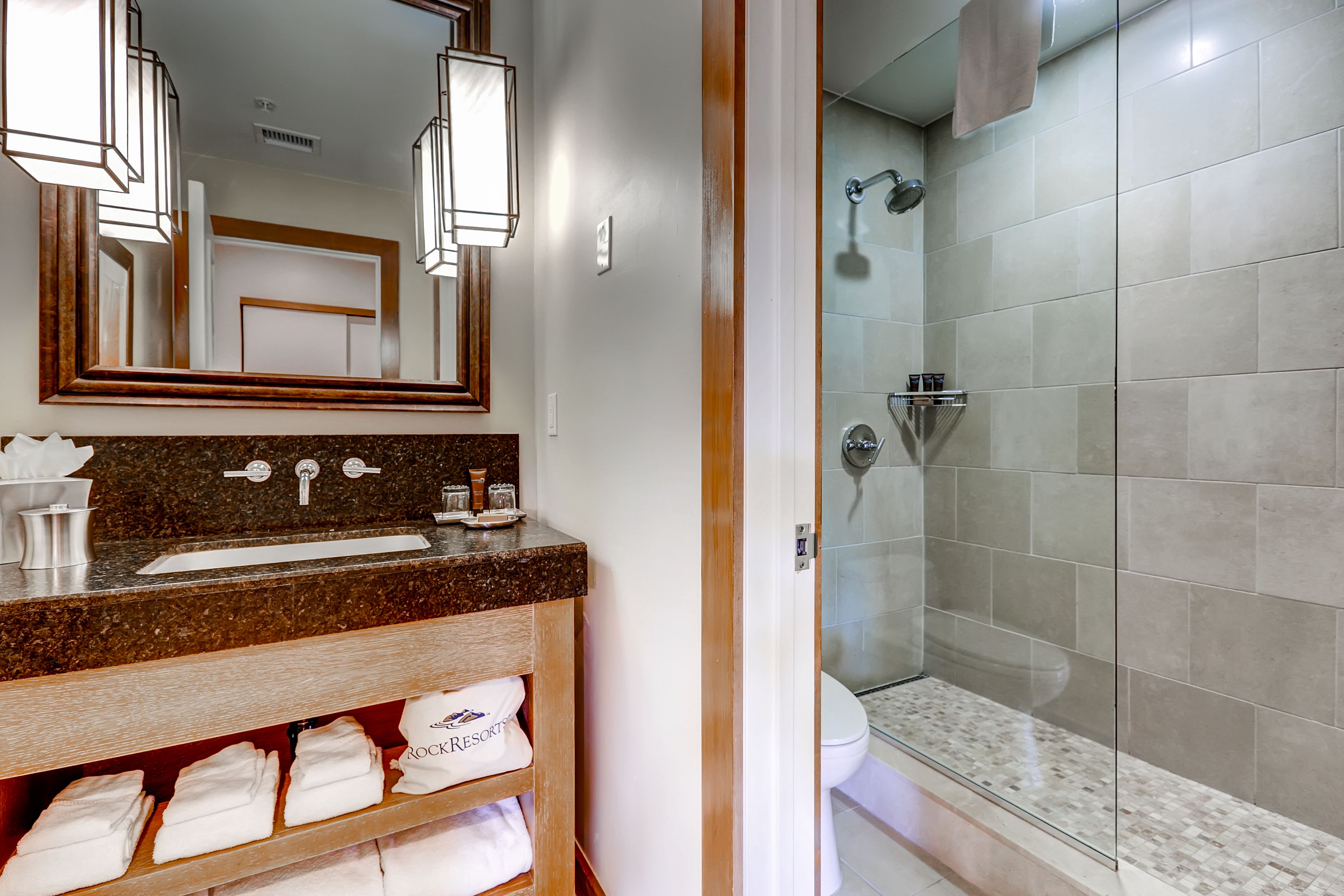 A bathroom with a granite countertop sink, a mirror with a wooden frame, a towel rack with white towels, and a glass-enclosed shower area.