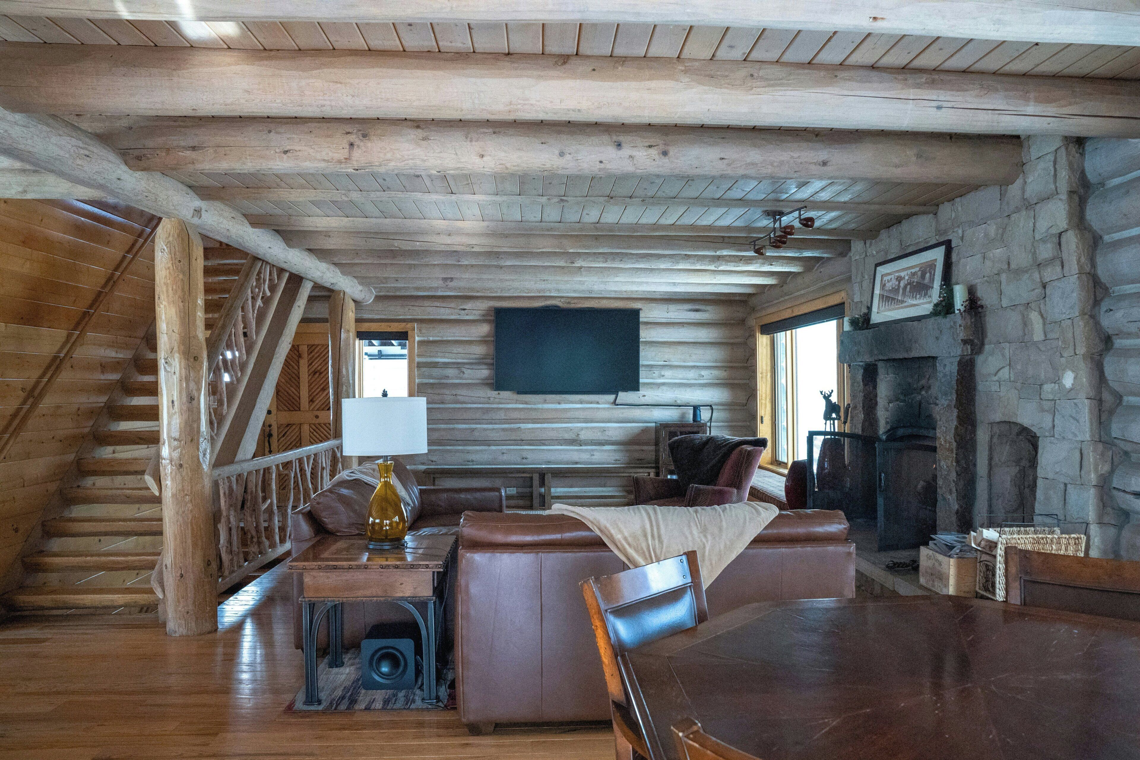 A log cabin interior with a wooden staircase, a fireplace, a television mounted on the wall, and a seating area with a table and chairs.