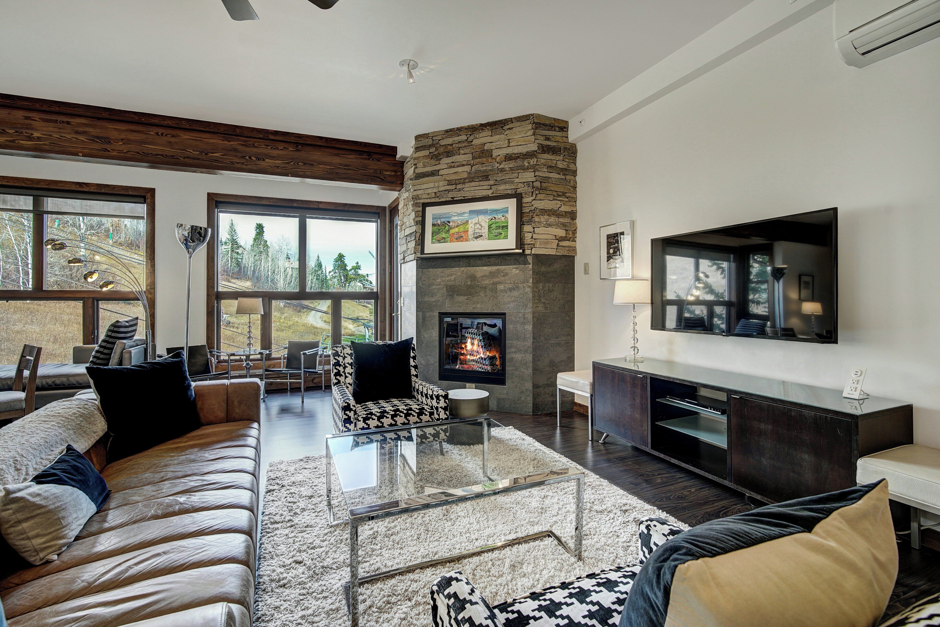 Un salón moderno con chimenea de piedra, sofá de cuero y amplios ventanales con vistas al exterior.