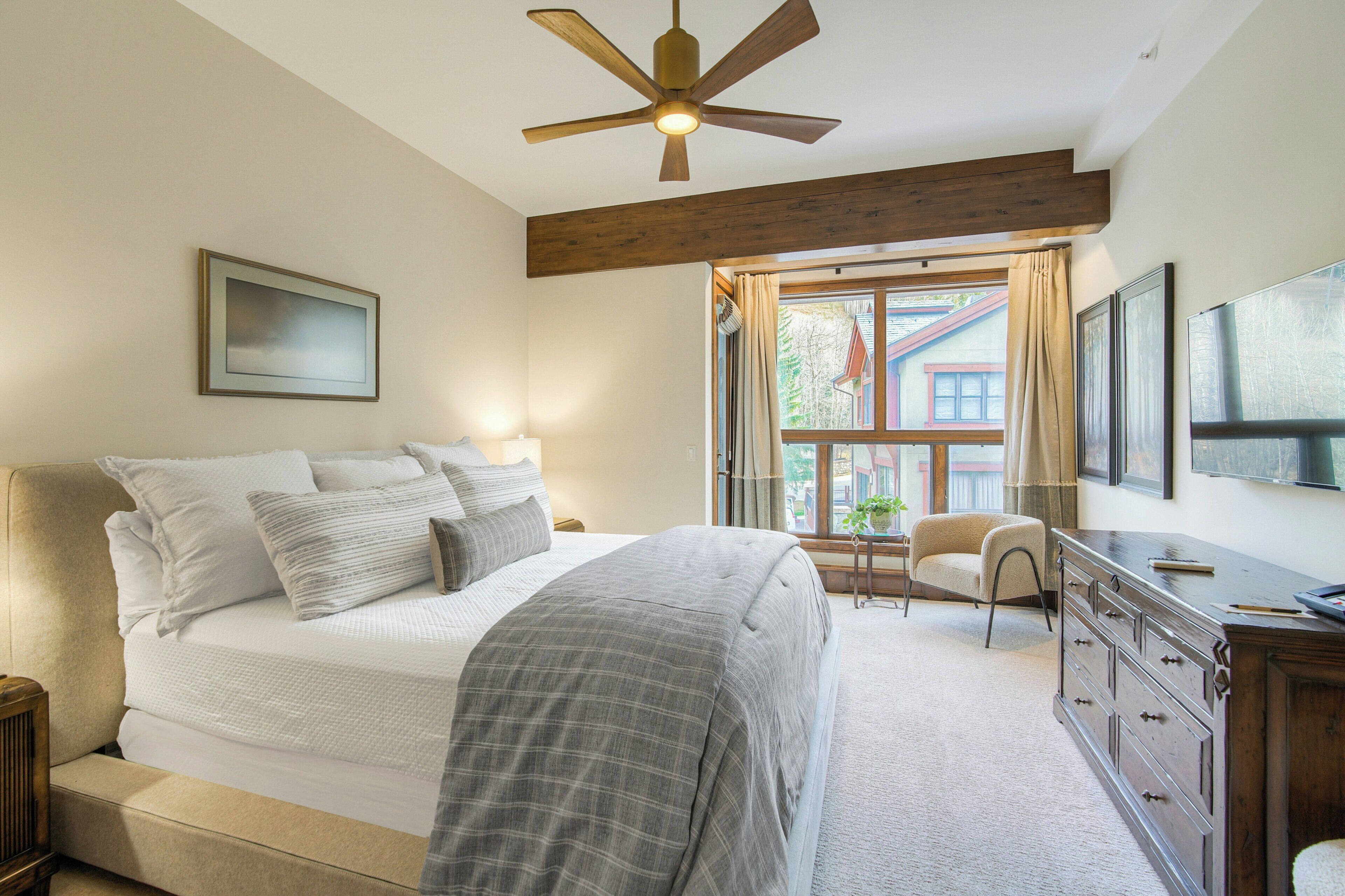 A bedroom with a large bed, wooden beams, a ceiling fan, a dresser, and a window with curtains.